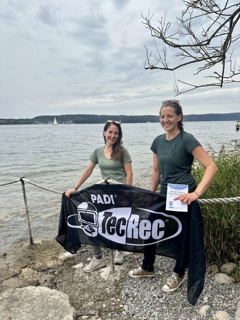 Two smiling women stand on a rocky lakeshore, holding a large PADI TecRec banner with sailboats on the lake in the background under a cloudy sky.