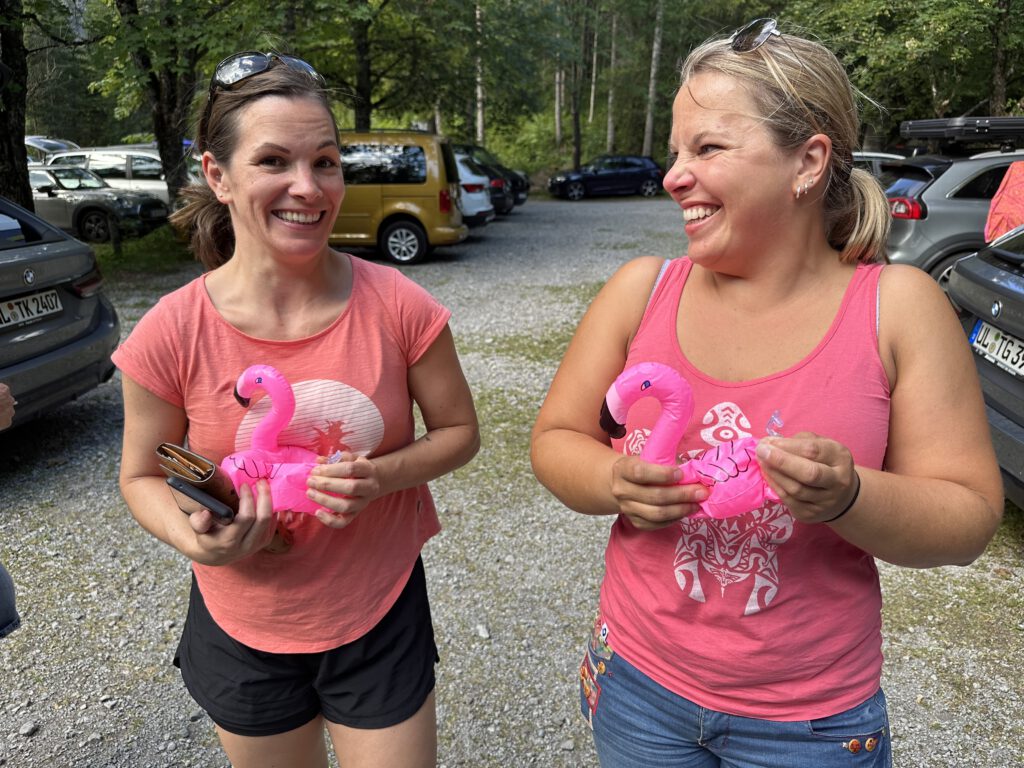 Two women smiling and talking outdoors, each holding a pink inflatable flamingo in a gravel parking lot with trees behind them.
