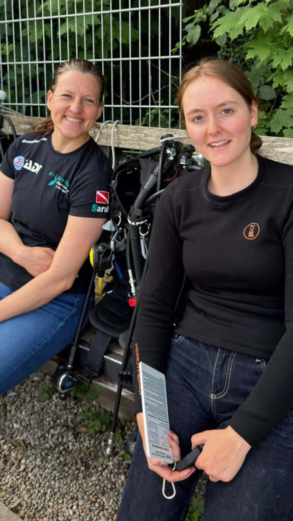 Two divers sit on a bench with scuba gear, smiling at the camera, greenery and a metal fence in the background.