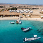 Turquoise sea at a beach resort with a wooden pier, boats, and white cabanas along a sandy shore.